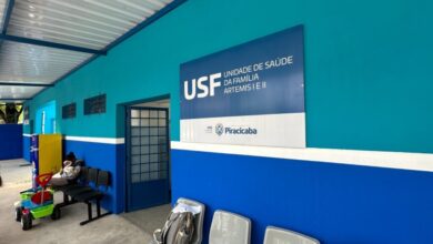 A imagem mostra a fachada de uma Unidade de Saúde da Família (USF) com a identificação "USF UNIDADE DE SAÚDE DA FAMÍLIA ARTEMIS I E II". A parede principal é pintada em um tom vibrante de azul-petróleo, com uma faixa branca separando-a da parte inferior, que é pintada de azul royal. À esquerda, há uma área coberta com um telhado de metal aparente, sustentado por vigas azuis. Nessa área, vemos alguns elementos, como uma porta de vidro com grades, duas caixas pretas retangulares fixadas na parede, um refrigerador azul com detalhes amarelos e um carrinho de brinquedo colorido. Ao lado, há um banco com algumas pessoas sentadas, uma delas segurando um objeto branco. Na parede principal, um grande painel azul exibe o nome da unidade de saúde e os logotipos da SMS (Secretaria Municipal de Saúde) e da Prefeitura de Piracicaba. Mais à direita, vislumbramos parte de uma entrada com porta de vidro e grades, e alguns assentos cinza, um deles com uma mochila sobre ele. A iluminação sugere que a foto foi tirada durante o dia, com luz natural entrando pela área coberta.